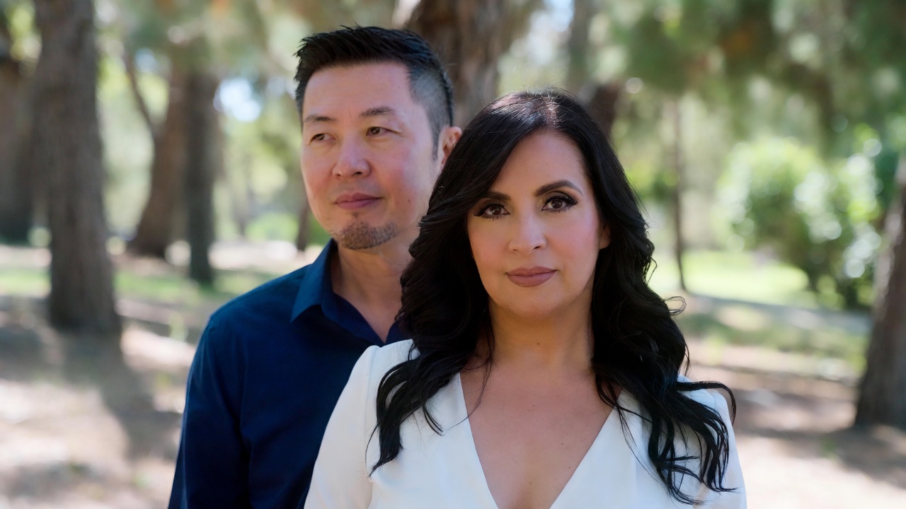 Asian man with short styled black hair and goatee standing beside a confident Asian woman with long dark wavy hair in a white V-neck top, both posing in a sunlit park